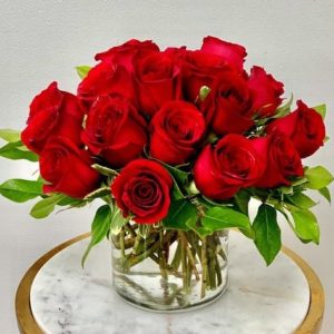 Twenty premium red roses compactly arranged in a 5" cylinder vase with lush greenery.