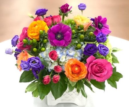 A vibrant combination of hot pink, orange, blue and purple flowers. This floral arrangement is designed in a white ceramic lattice box. 